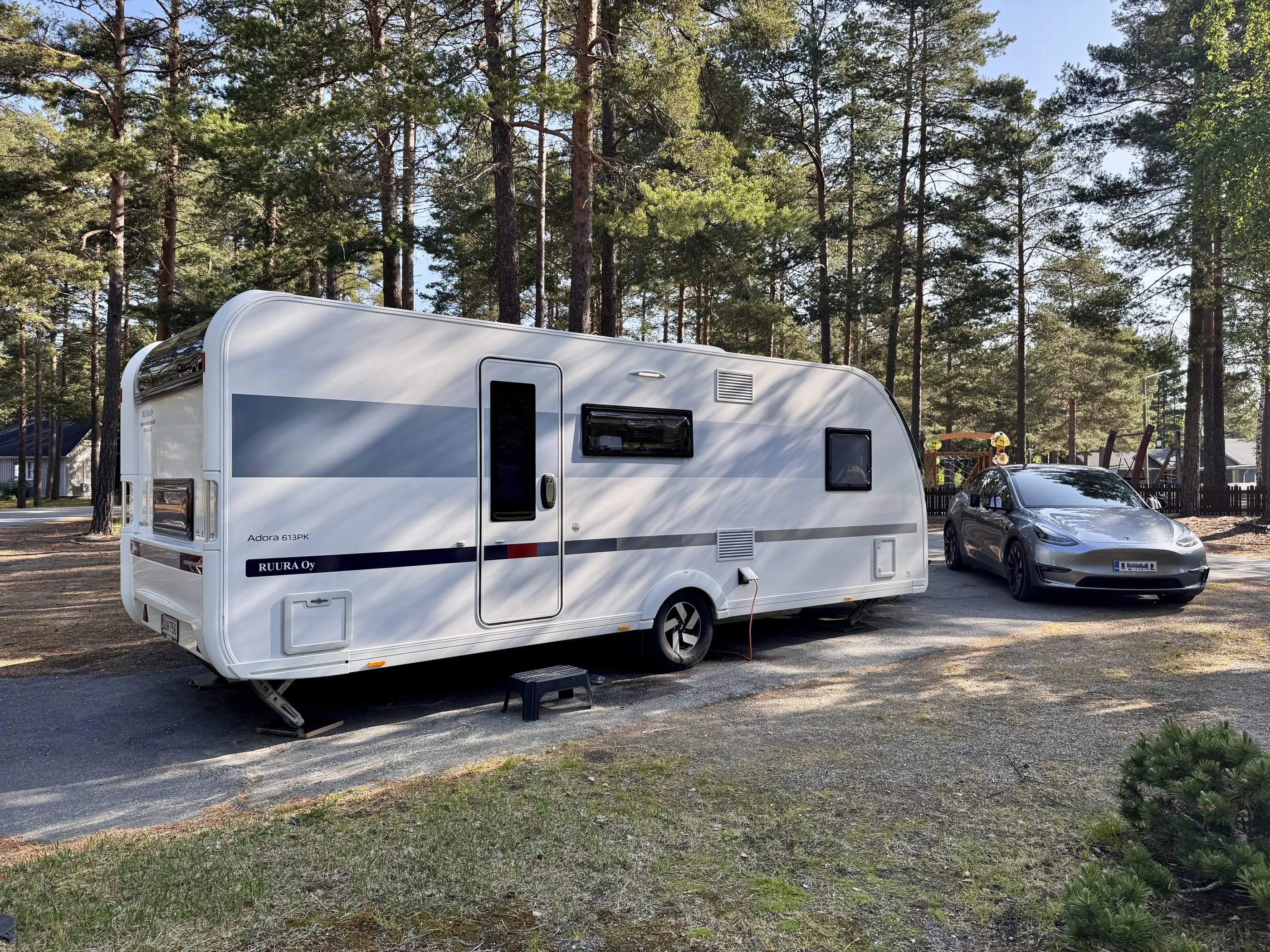 Kuva esittää valkoista asuntovaunua ja harmaata Teslaa metsäisellä alueella aurinkoisena päivänä.