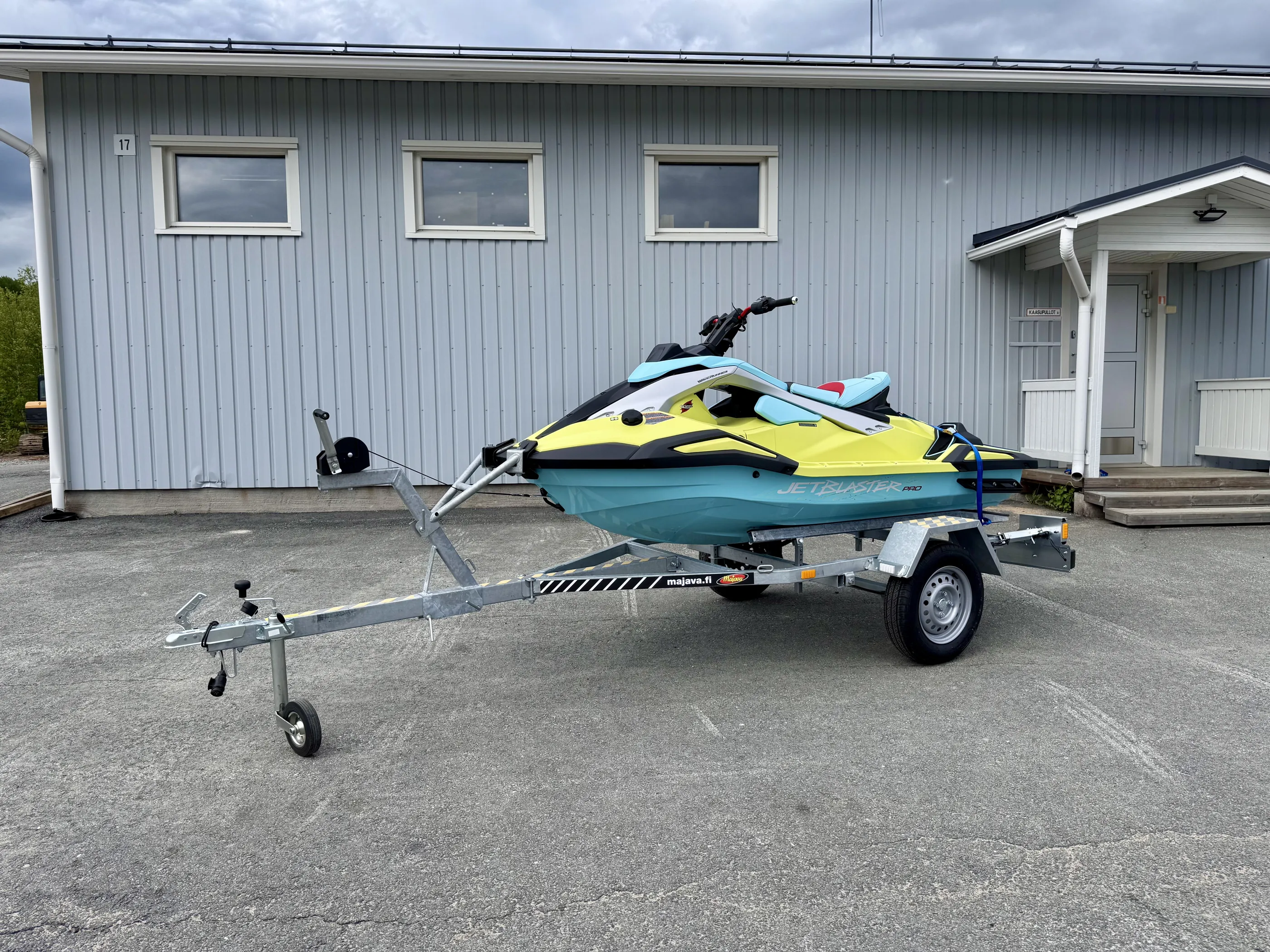 Vaaleansininen ja keltainen vesiskootteri trailerilla harmaalla pihalla, rakennus taustalla, pilvinen sää.